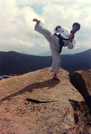 Master Han Kyu Yoon performing a side kick atop a Colorado mountain boulder with the Rocky Mountains behind him